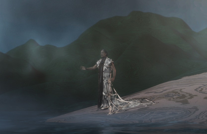 A person stands on a shore with an arm outstretched and mountains behind them