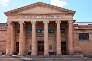 A sandstone building with Art Gallery of New South Wales written at the front above columns, and a panel featuring hooded heads above the door