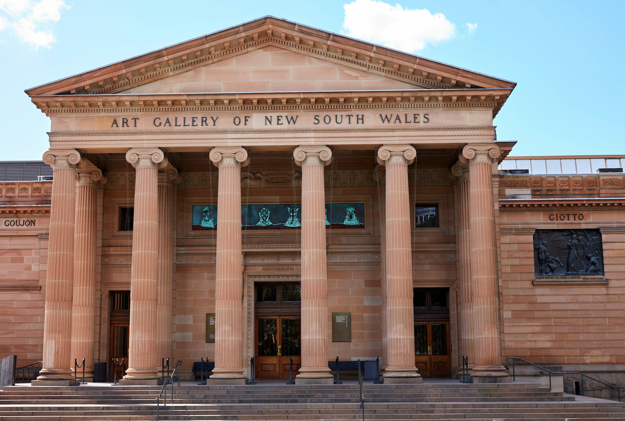 A sandstone building with Art Gallery of New South Wales written at the front above columns, and a panel featuring hooded heads above the door