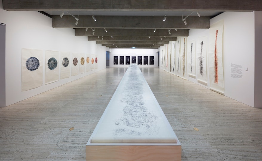 A gallery space with artworks hung on the walls and an extremely long drawing on paper on a central plinth
