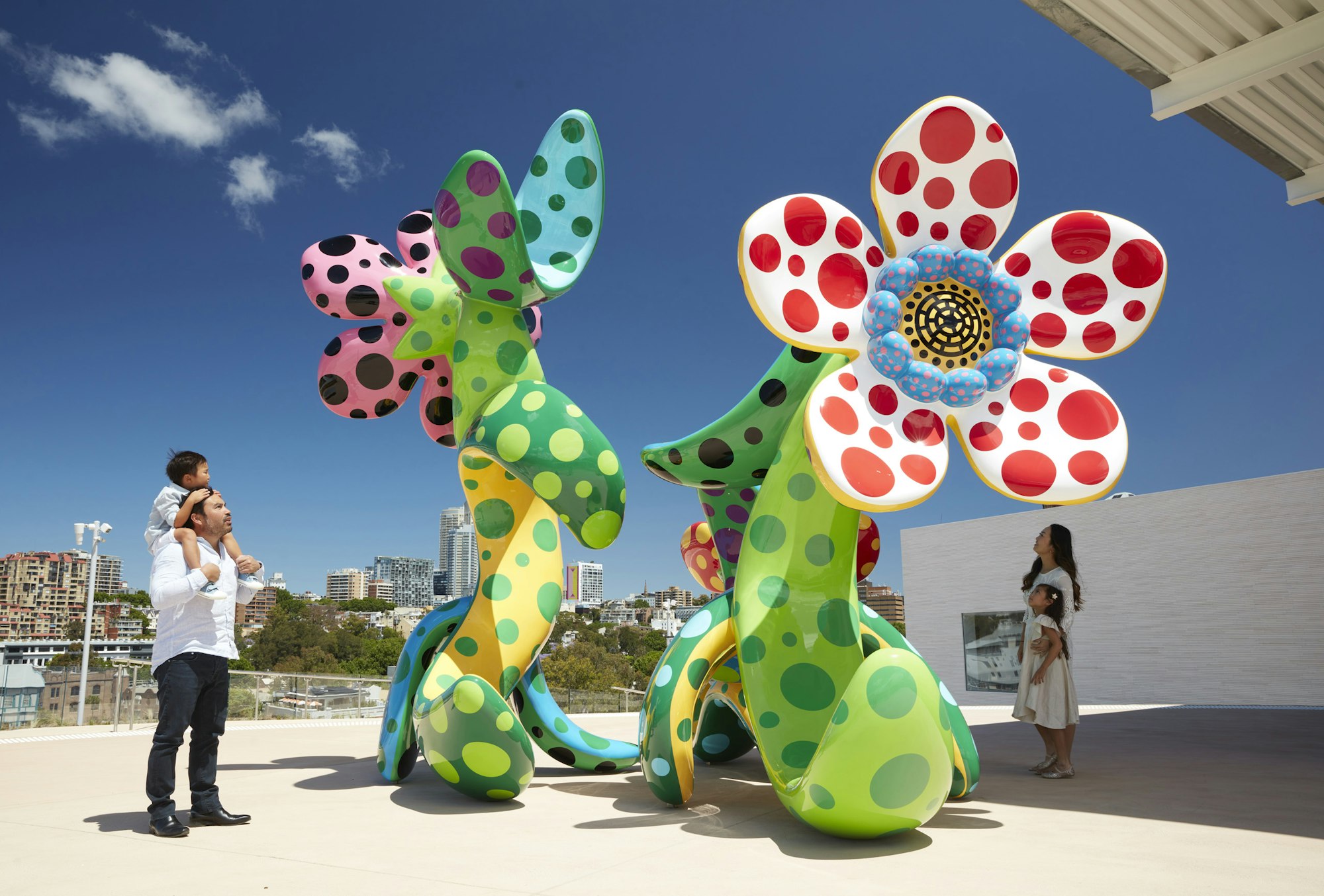 Four people look at a colourful sculpture of flowers with spots on an outdoor terrace