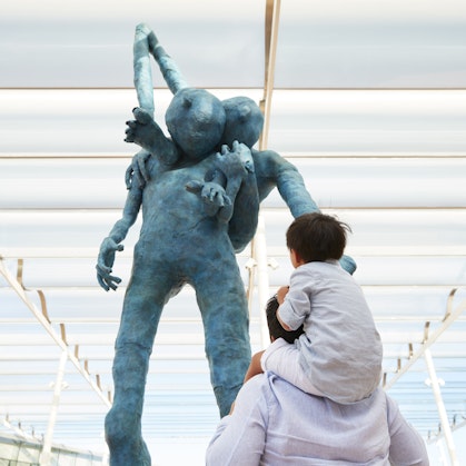 A child on an adult's shoulders looks up at a big blue figure carrying another on their back