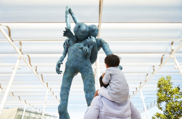 A child on an adult's shoulders looks up at a big blue figure carrying another on their back