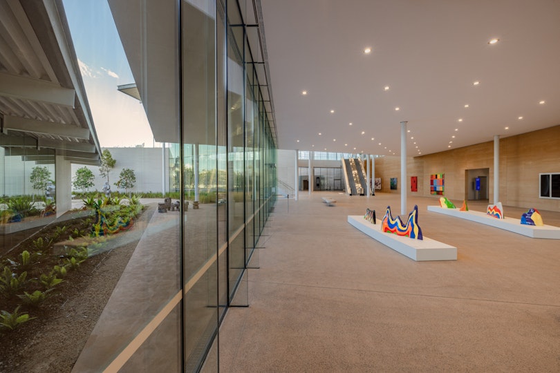 On left an outdoor garden with sculptures. On right, through a glass wall, a gallery space with artworks on plinths and on an earthen wall