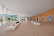 A large gallery space with a glass wall on left, earthen wall on right featuring artworks on the walls and on plinths