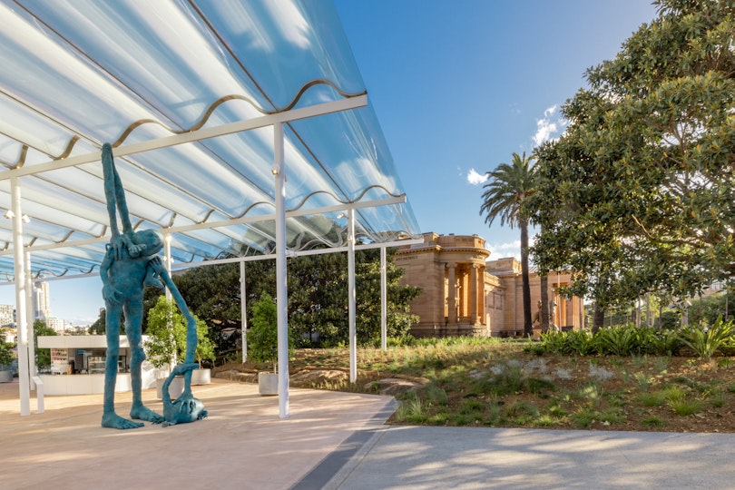 A large figurative sculpture under an awning with grass, trees and a building in the background