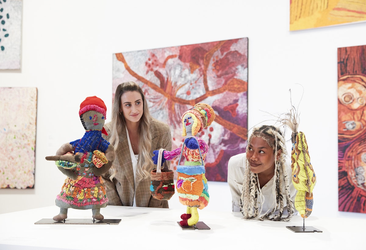 Two people looking at small figurative sculptures. Paintings hang on a wall behind them.