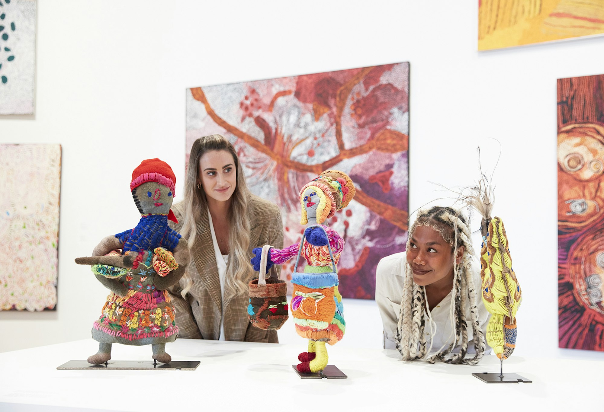 Two people looking at small figurative sculptures. Paintings hang on a wall behind them.