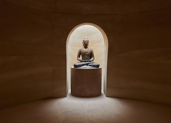 A seated Buddha sculpture on a plinth within a niche in earthen-walled room