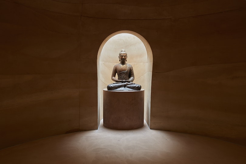A seated Buddha sculpture on a plinth within a niche in earthen-walled room