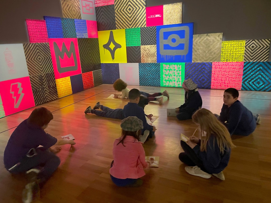 Seven children on the floor of a gallery space writing on paper with an artwork made up of colourful squares on two walls behind them