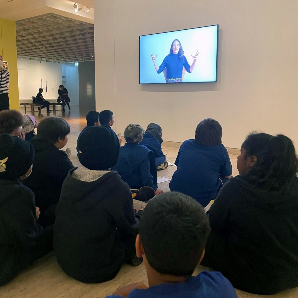 A group of children sit on the floor watching a person on a wall-mounted screen