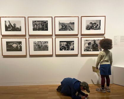 In a gallery space, one child looks at framed photographs on a wall while other kneels on the floor writing on paper