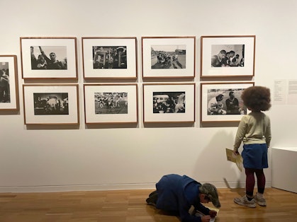 In a gallery space, one child looks at framed photographs on a wall while other kneels on the floor writing on paper