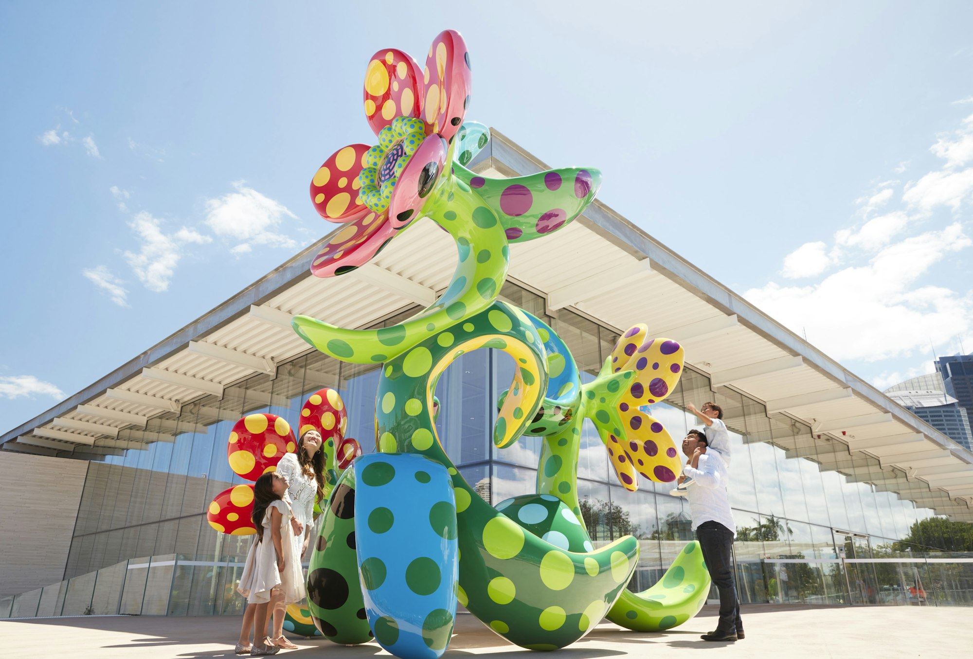 Two adults and two children look at a large sculpture of a colourful, spotted flower in front of a glass building