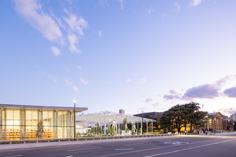 A glass-walled building on left and a sandstone building on right with a covered plaza and large tree in between