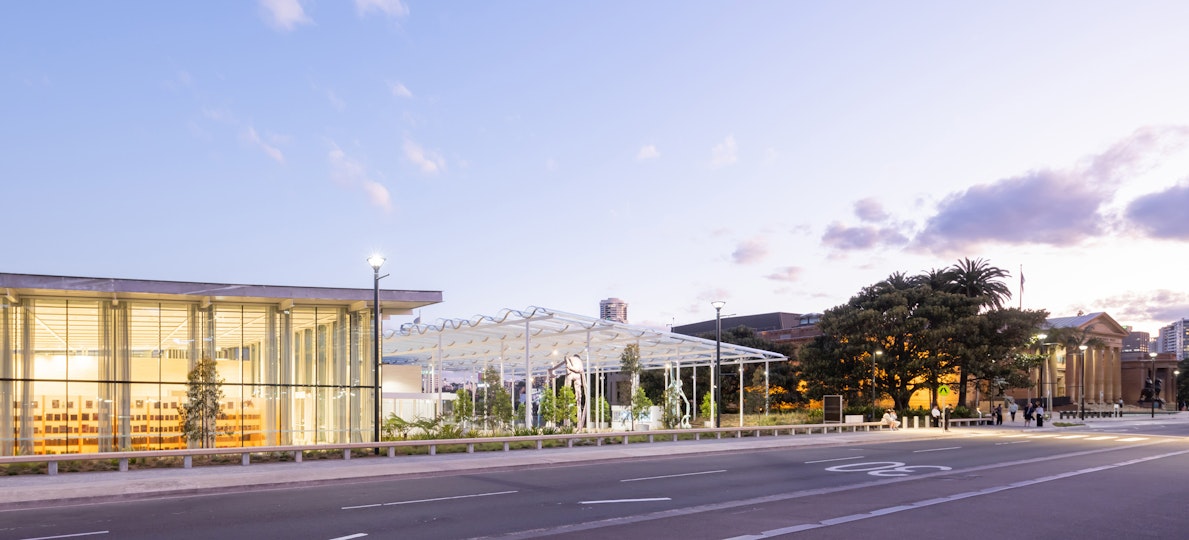 A glass-walled building at left, a sandstone building at right with a large tree and plaza with a roof in the middle