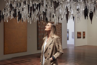 A person in a gallery space with paintings on the walls and an artwork of glass shards above them