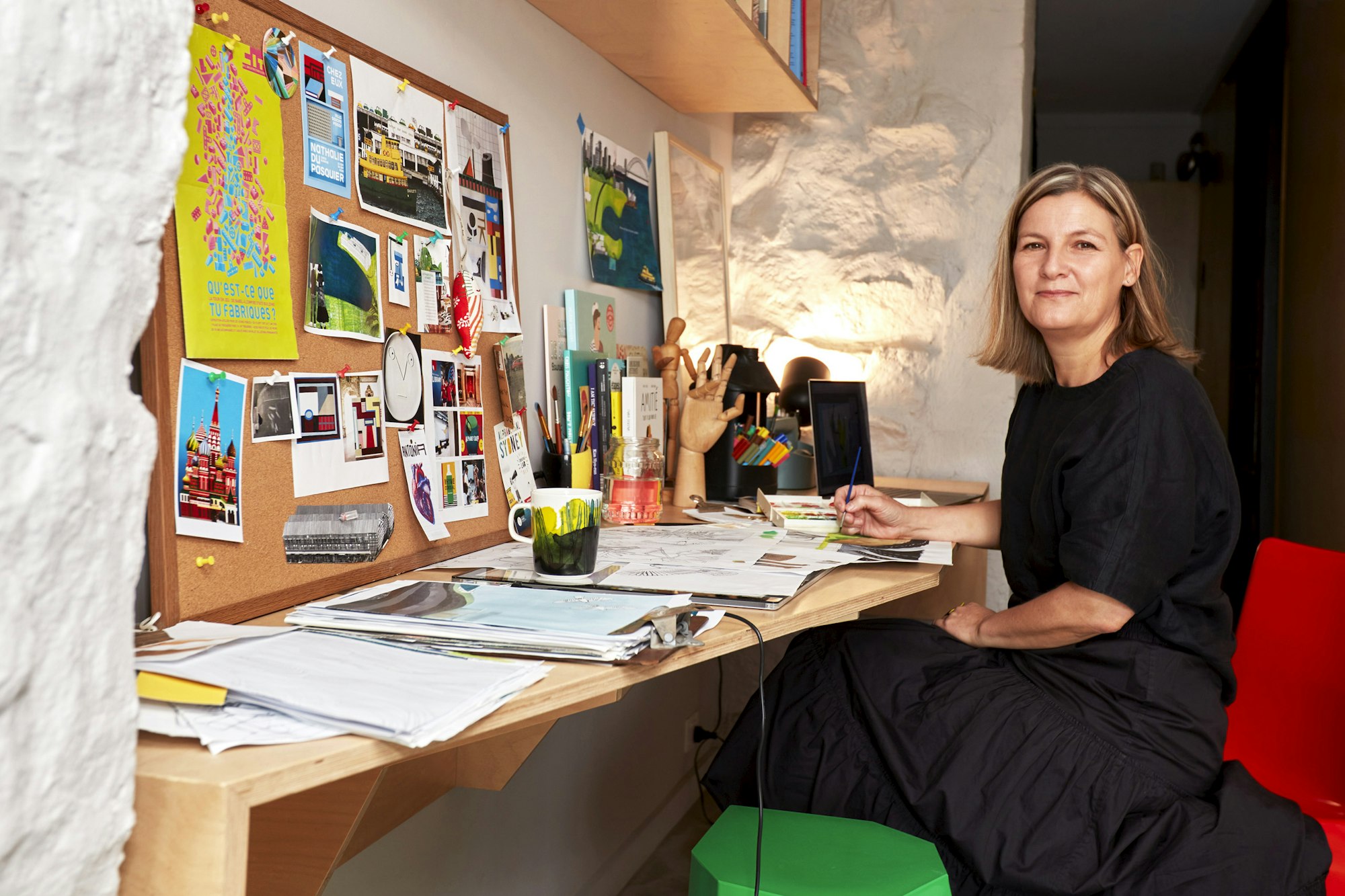 A person sits at a desk on which there are many items, including piles of paper and a pinboard