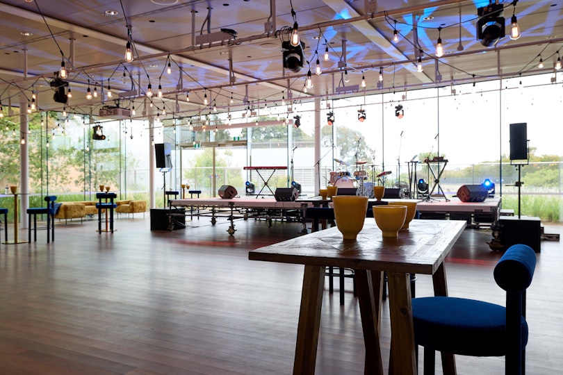 A glass-walled room with a few tables, stools and lounges, and a raised stage set up for a musical performance