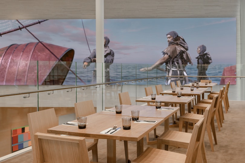 Dining tables and chairs in the foreground with a large artwork in the background depicting people on a vessel on water