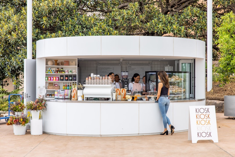 A person stands in front of staff at the counter of a small circular building with a sign that says kiosk