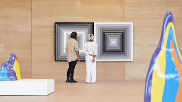 Two people in front of a work of art on a wall, with two colourful sculptures in the foreground