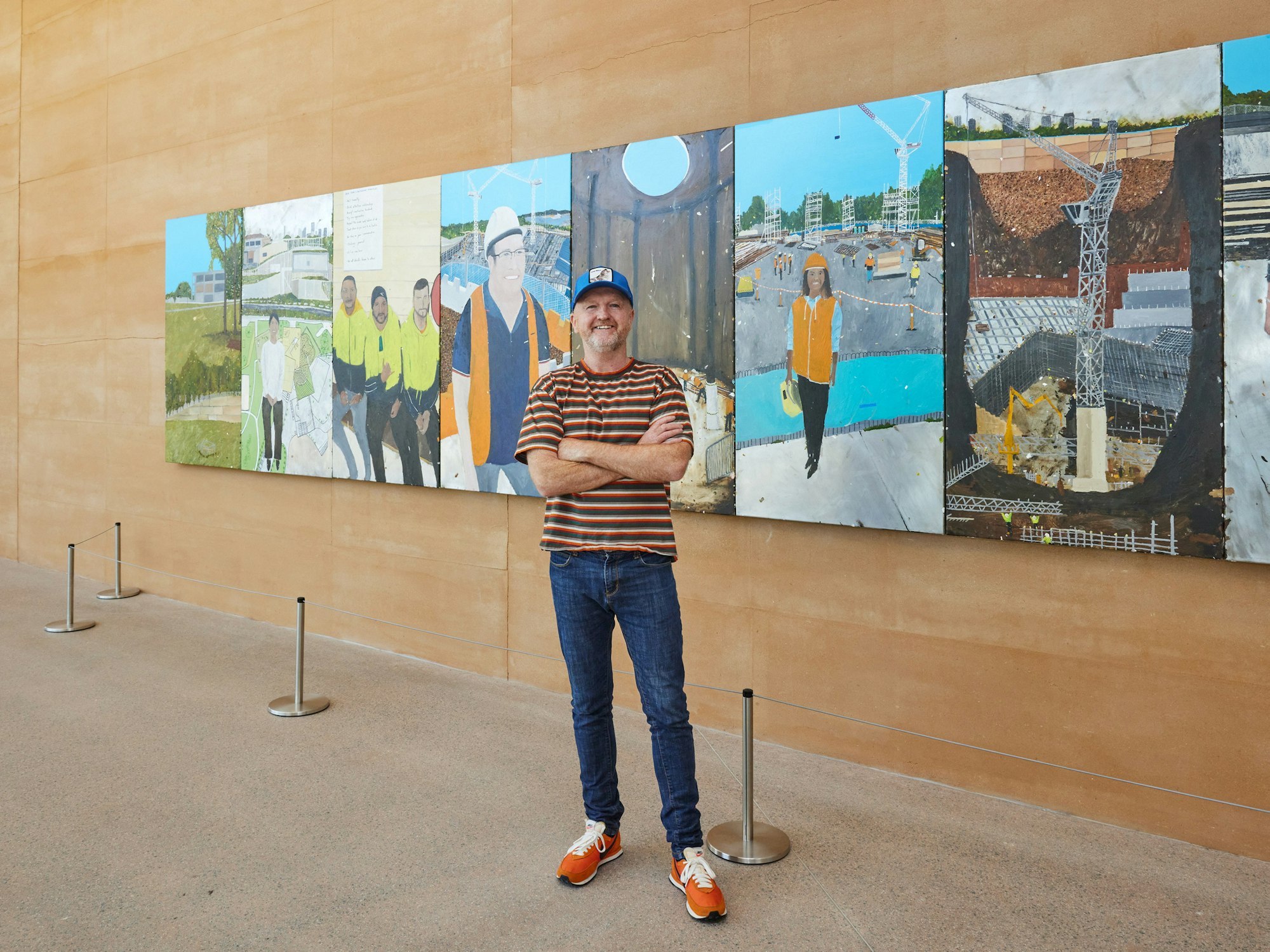 A person stands in front of a large multi-panelled painting of a construction site and various workers