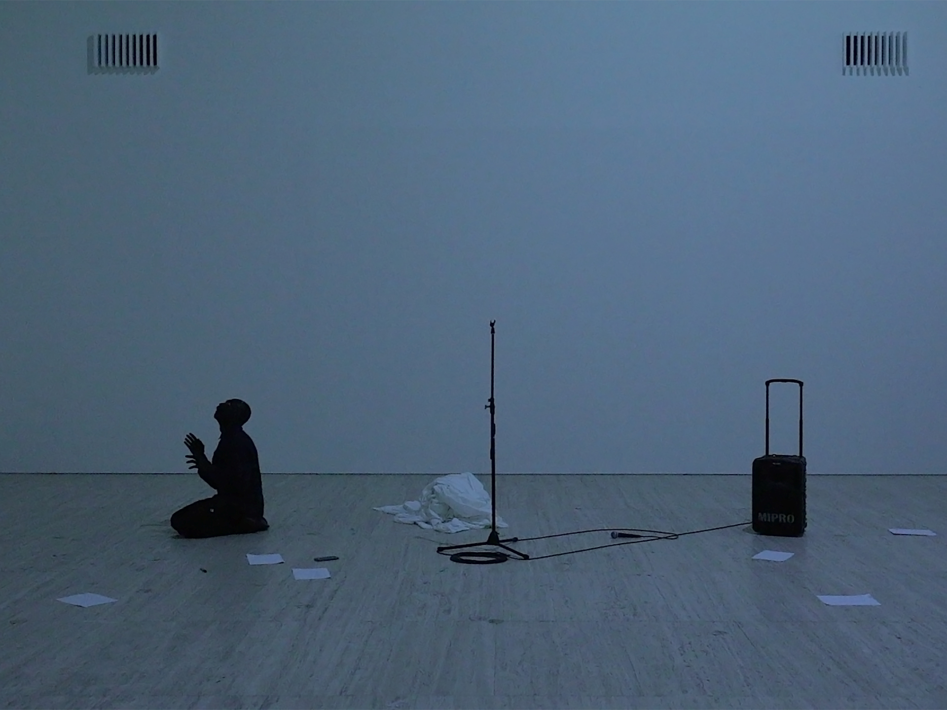 A person in kneeling with hands clasped, looking up in a dimly-lit room. Behind them is a microphone stand, a speaker and a crumpled sheet.