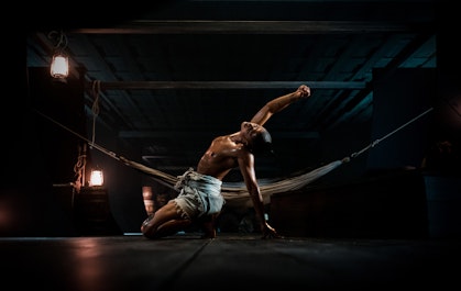 A squatting person with arms outstretched in a dark room with a hammock, lit by two hanging lamps