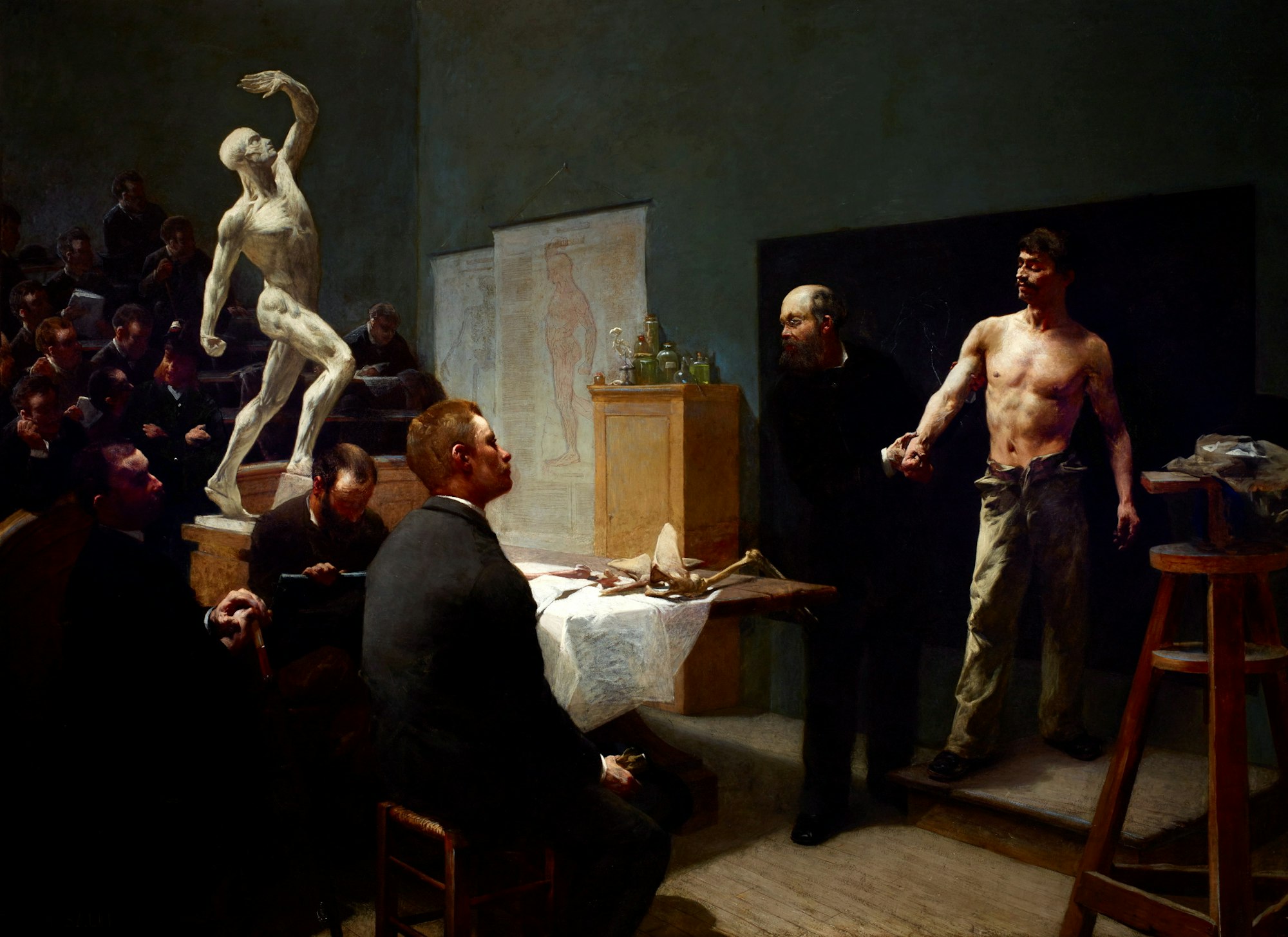 A scene at an anatomy class: the model with a bared torso has their hand held by the teacher. Adult students sit and look at the model.