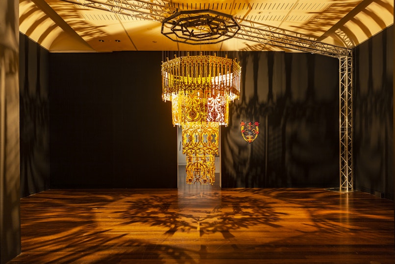 A large chandelier of intricate patterns and shapes that is lit up to cast dramatic shadows across the room. Behind the chandelier is a smaller artwork affixed to a wall.