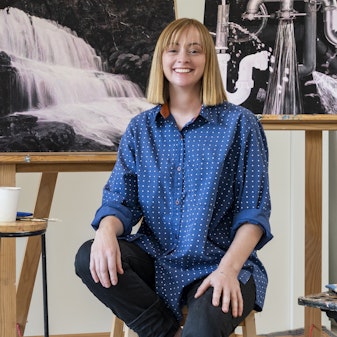 A person sits on a stool between artworks on easels