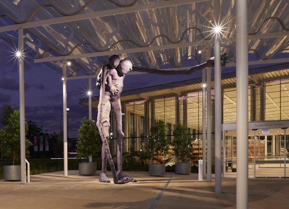 A large purple sculpture of a figure with another on its back, in front of a lit building at night