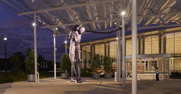 A large purple sculpture of a figure with another on its back, in front of a lit building at night