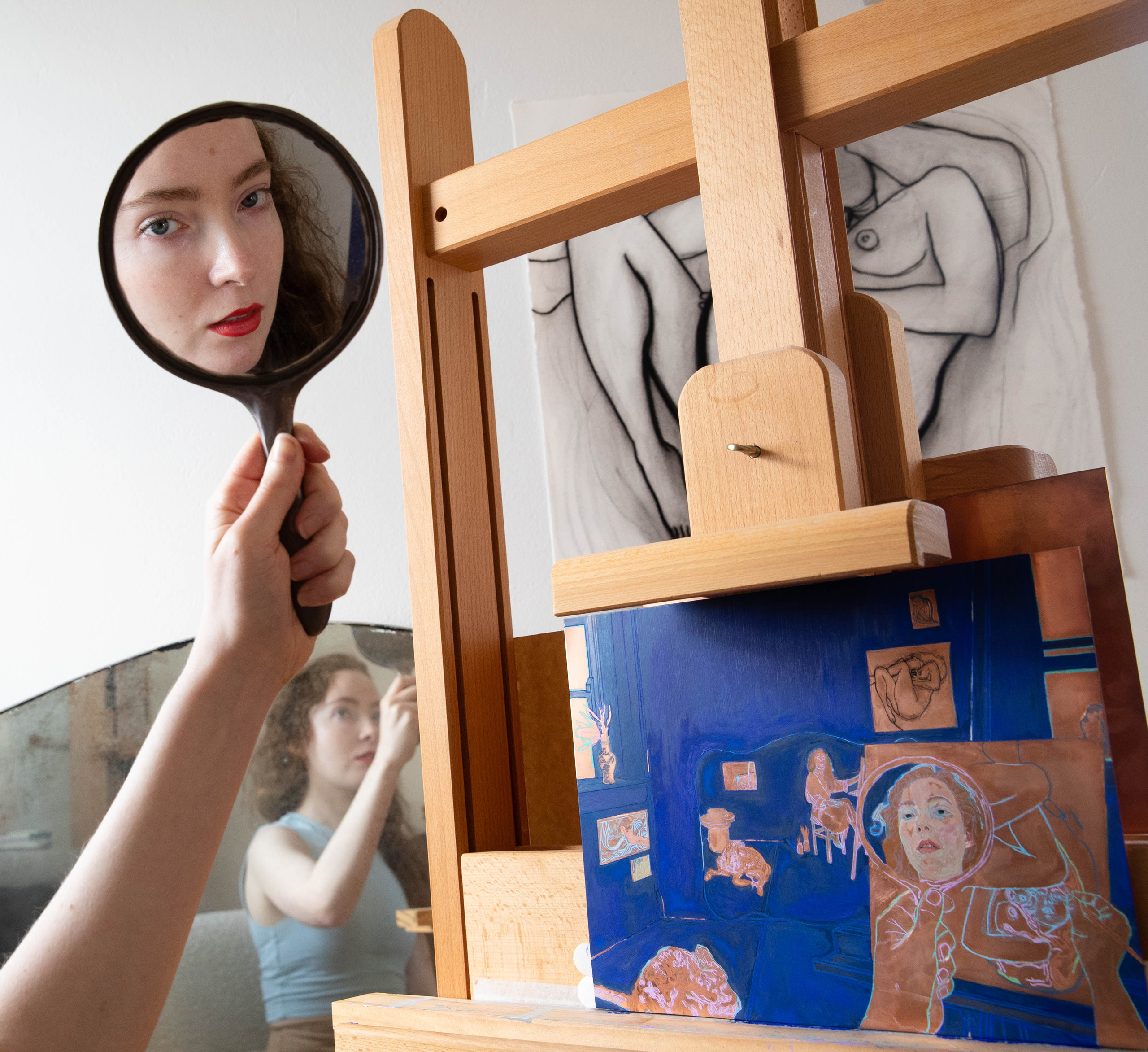 A hand holds a small mirror in which a face is reflected. Below more of the person is reflected in a larger mirror. To the right is an easel and artwork.