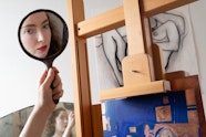 A hand holds a small mirror in which a face is reflected. Below more of the person is reflected in a larger mirror. To the right is an easel and artwork.
