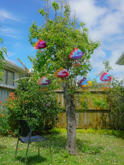 A photograph of tree in a backyard that is hung with six colourful, cloud shapes with long tassles. Facing the tree is a black chair.