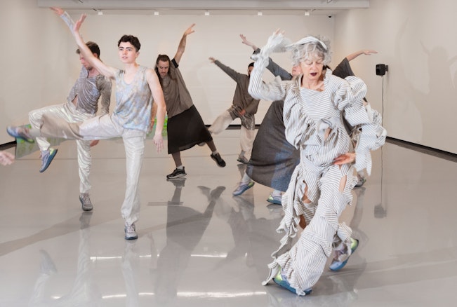A group of 6 people dancing in a large white room. All of them are dressed in silver or grey costumes.