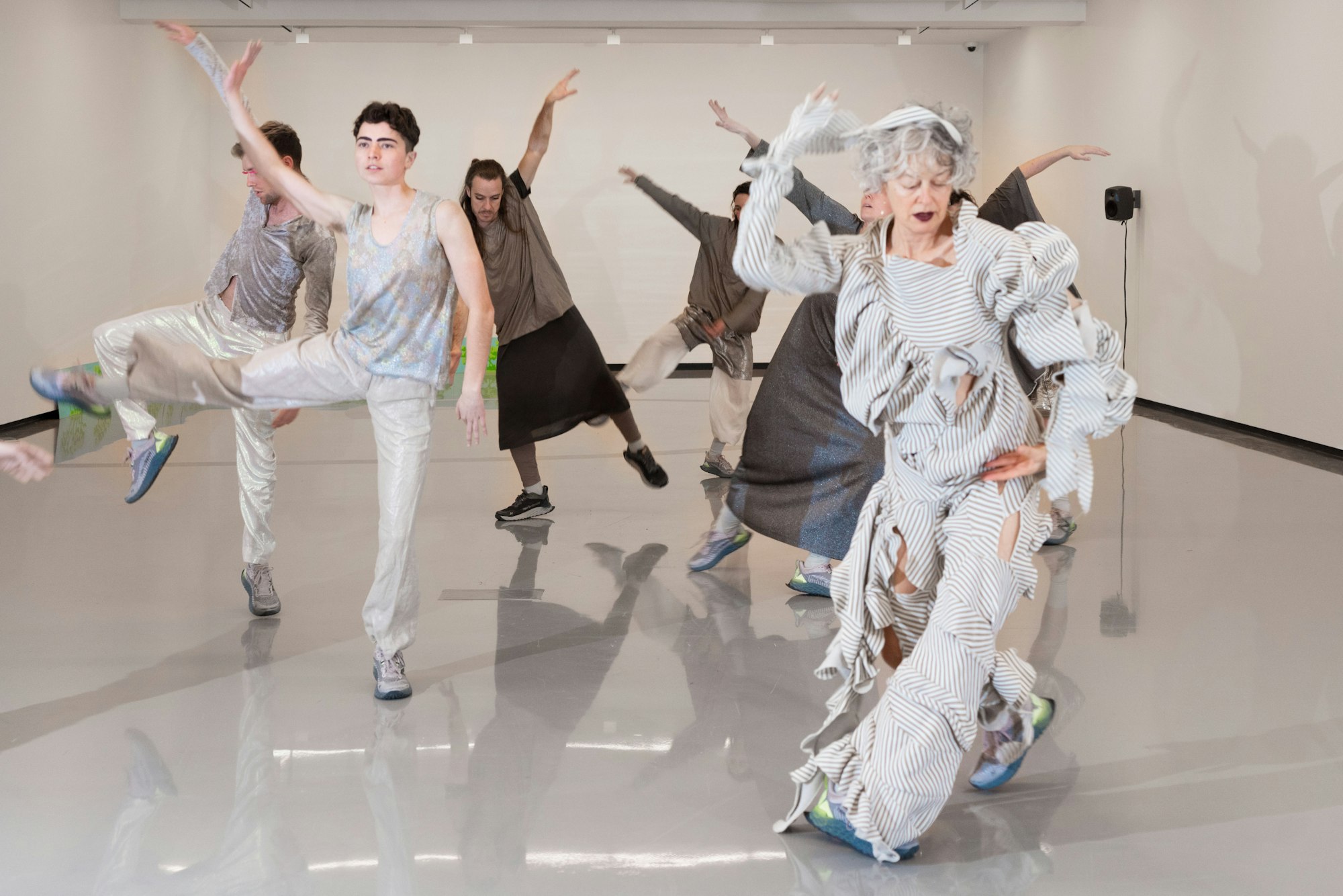 A group of 6 people dancing in a large white room. All of them are dressed in silver or grey costumes.