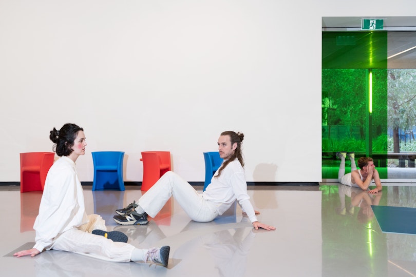 Two dancers in white costumes sitting opposite each other. Both have light skin and long brown hair. A third dancer is lying on their stomach in the background.