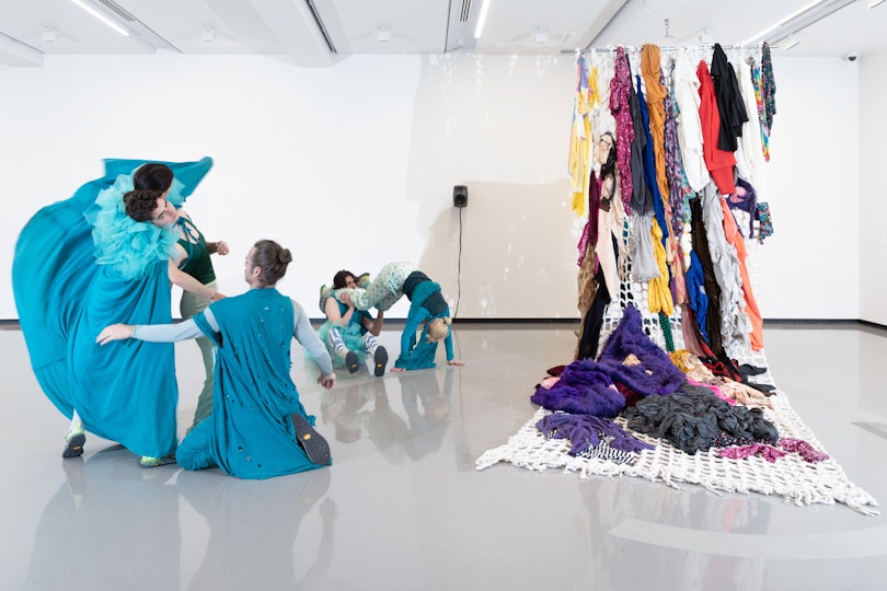 Five danceres in blue costumes moving in two groups in a white room. On the right are several costumes hanging from ceiling to floor.