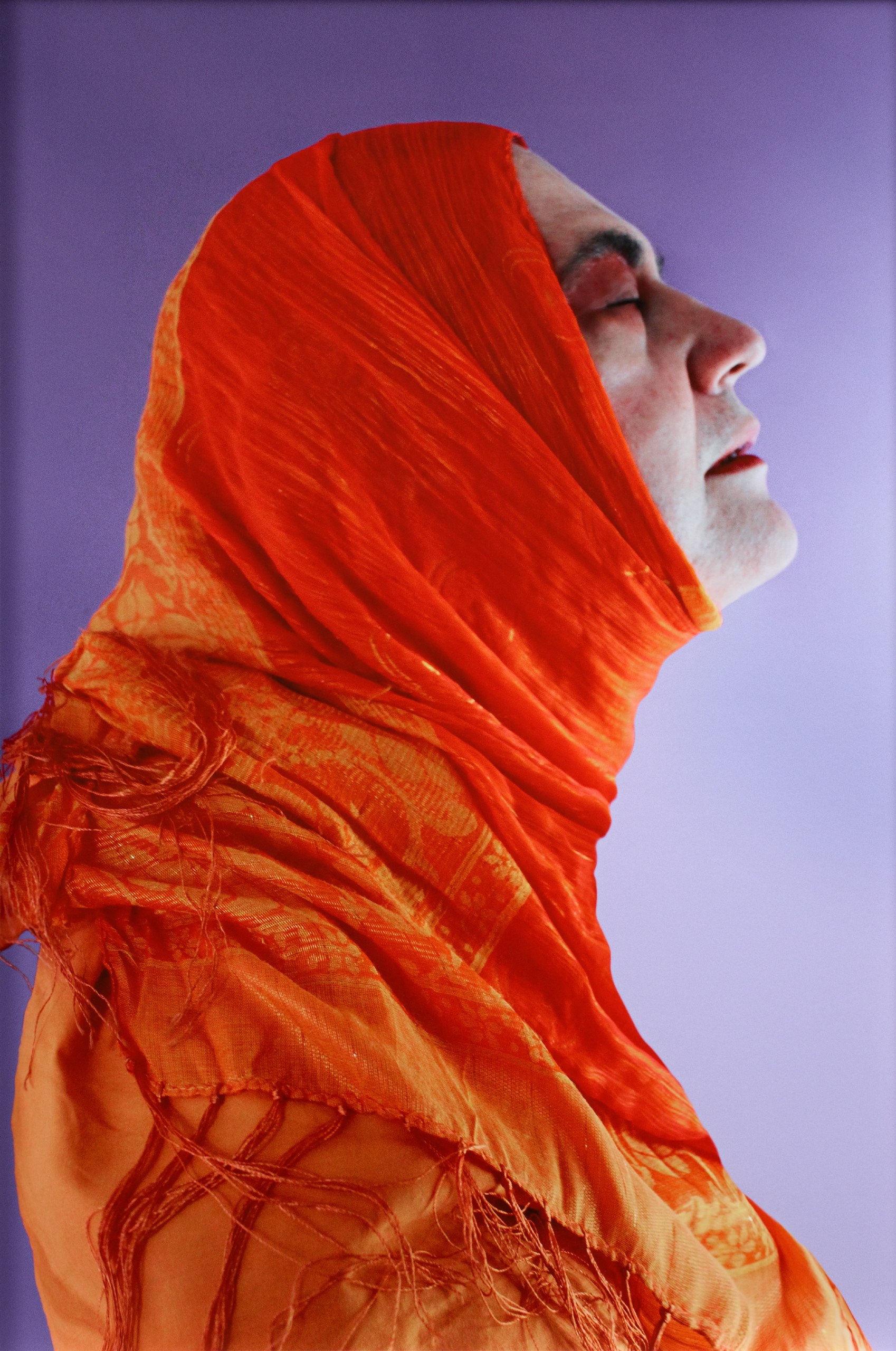 A photograph of the side profile of a person with closed eyes and their face tilted up. They are wrapped from head to torso in orange fabric and in front of a purple background.