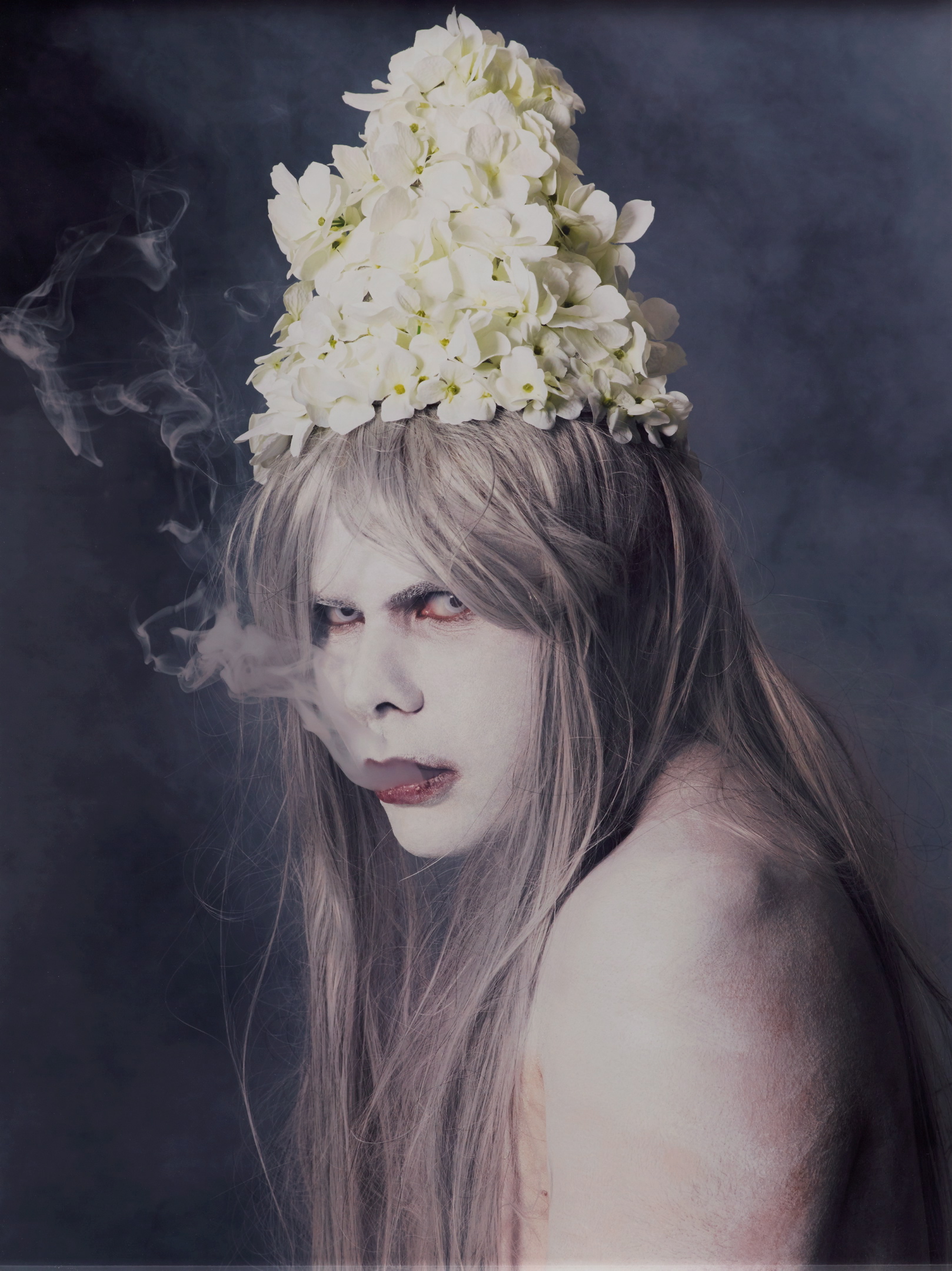 A photograph of a person with long blonde hair and pale skin looking into the camera. Smoke comes out of their mouth. On their head is a floral headpiece.