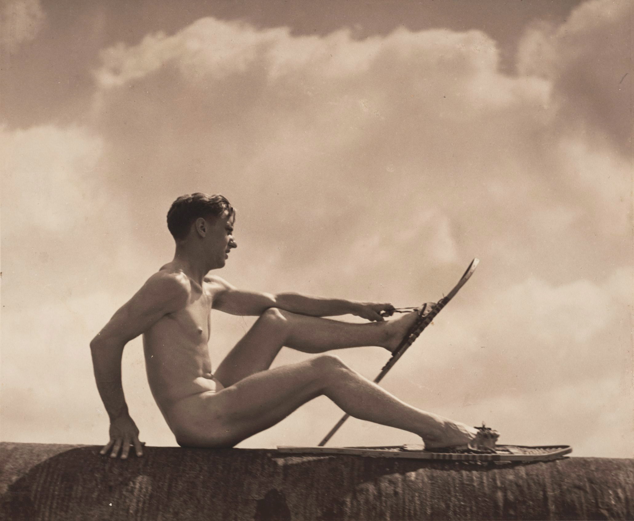 A seated nude person wearing snowshoes, viewed from the side. Behind them are clouds.