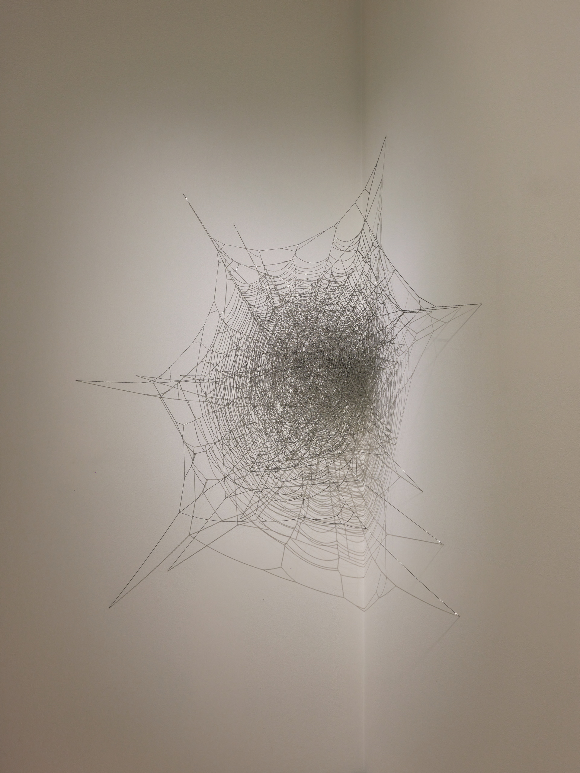 A sculpture of metal chains and. pins in the form of a dense spider web installed in a wall corner.