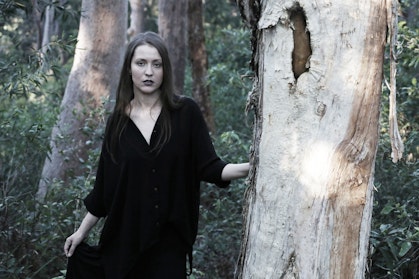 A person dressed in black with long hair stands in bushland next to a tree