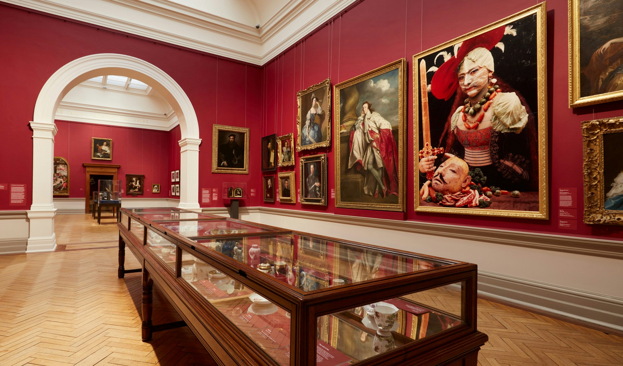 A red-walled gallery space with gold-framed paintings on the wall and a wood and glass cabinet with objects on the floor