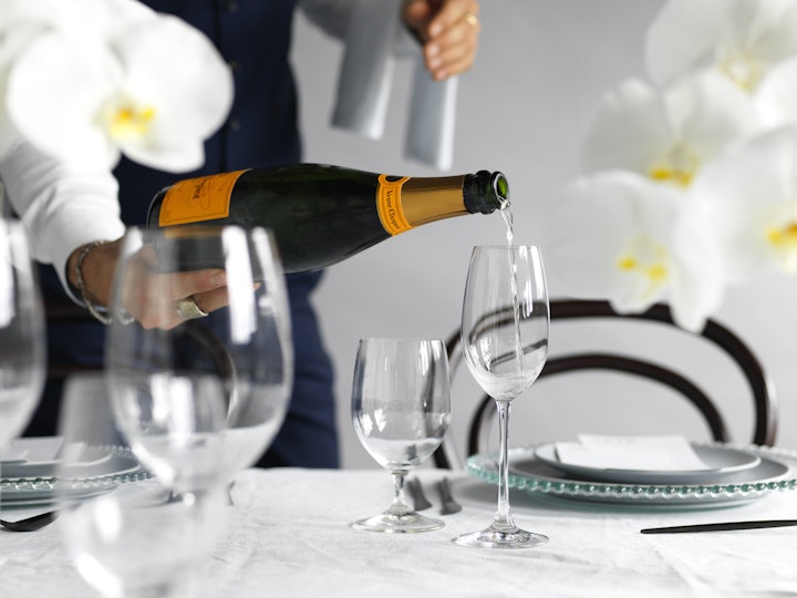A person pours champagne into a glass at a table set for a meal