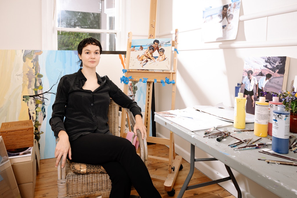 A person sits in a chair next to an artwork on an easel and a table holding other art and art materials